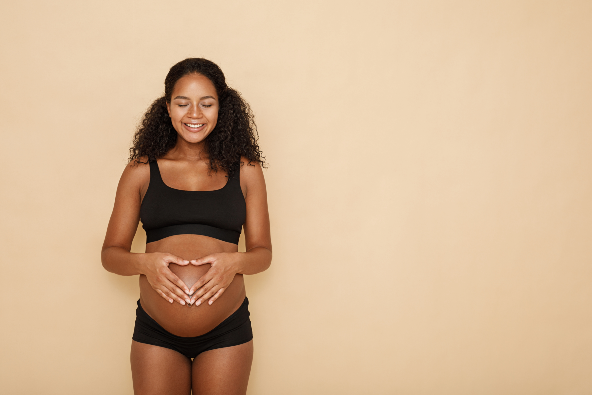 Pregnant woman with curly hair in black underwear, eyes closed, smiling, forms heart on belly with hands against beige background, expressing joy.