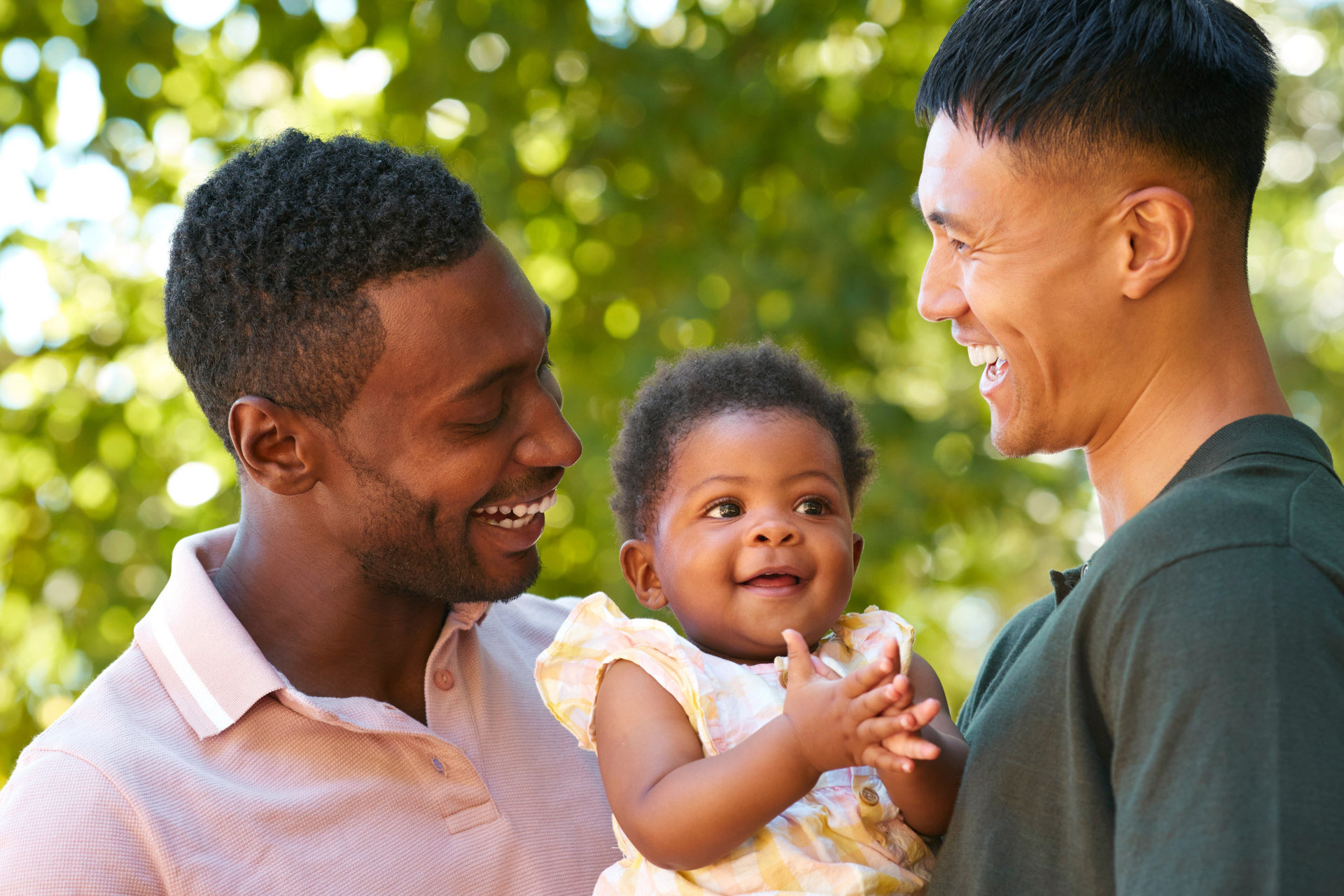 A happy family of two men holding a smiling baby outdoors. The men are looking lovingly at the child. Sunlight filters through green leaves in the background.
