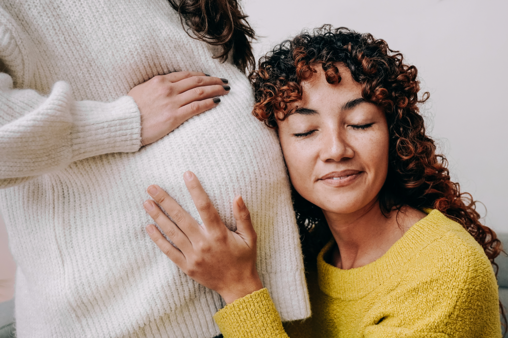 A woman in a yellow sweater leans her head joyfully against a pregnant belly, eyes closed. Another hand rests on the belly, creating a warm, serene scene.