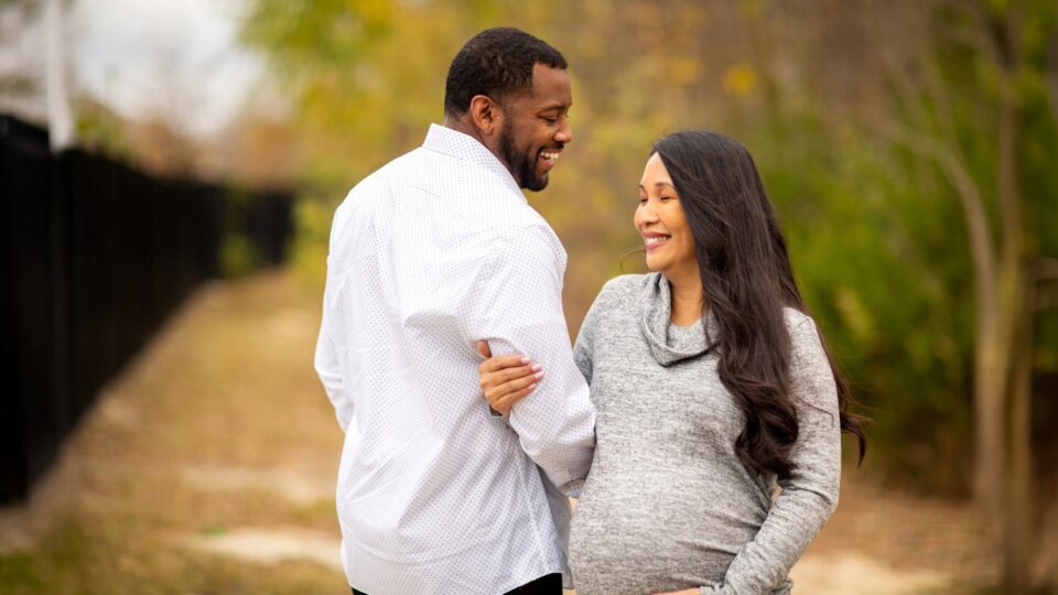 A couple stands outdoors, smiling warmly at each other. The woman is pregnant, wearing a gray sweater, holding the man's arm, with an autumn background.