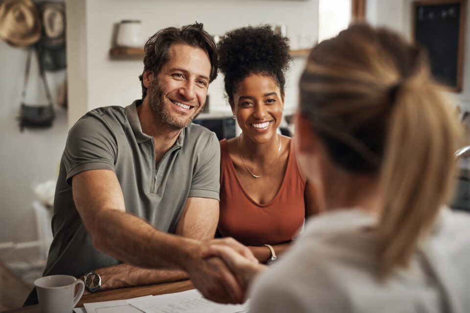 A smiling couple sits at a table, shaking hands with a person whose back is to the camera, conveying a friendly, welcoming atmosphere.