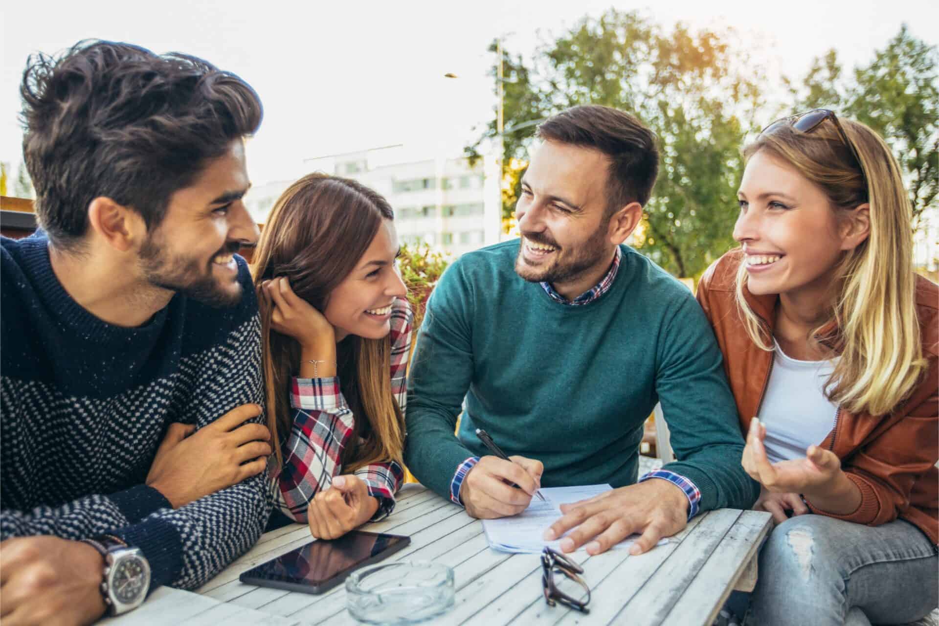 Four friends are sitting at an outdoor table, laughing and engaging in conversation. A man is writing on paper, while a tablet and eyeglasses are on the table.
