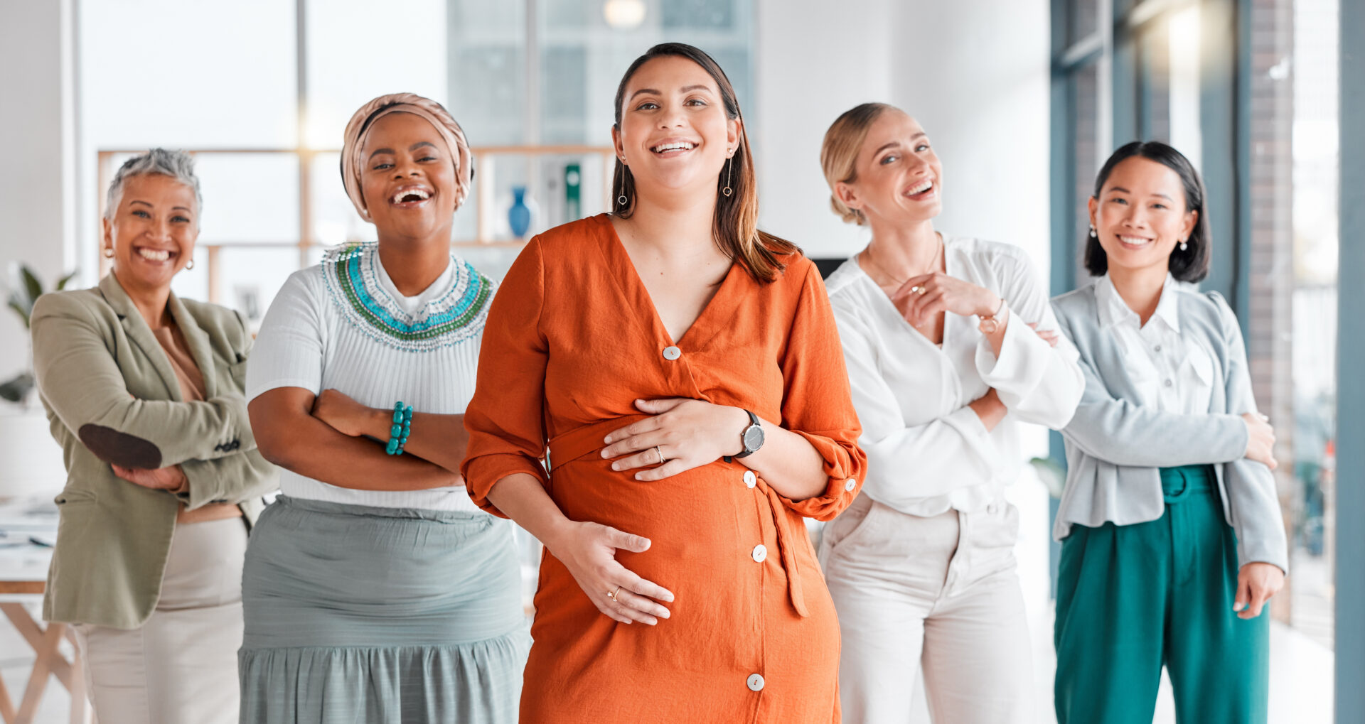 A diverse group of five smiling women in a brightly lit office. The woman in front, wearing an orange dress, appears pregnant. They exude confidence and joy.