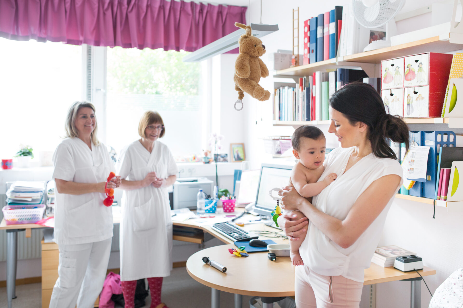 A smiling woman gently holds a baby in a bright office, with two women in white coats watching. The room is decorated with toys and books, conveying a cheerful, welcoming atmosphere.