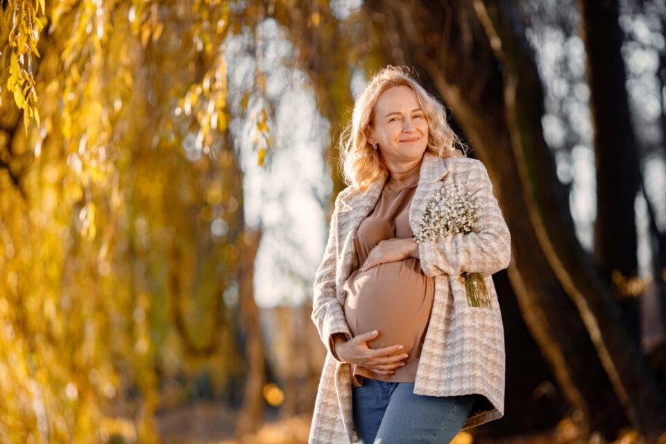A smiling pregnant woman stands outdoors in a sunlit autumn setting, holding her belly and a small bouquet of white flowers. Warm, joyful atmosphere.