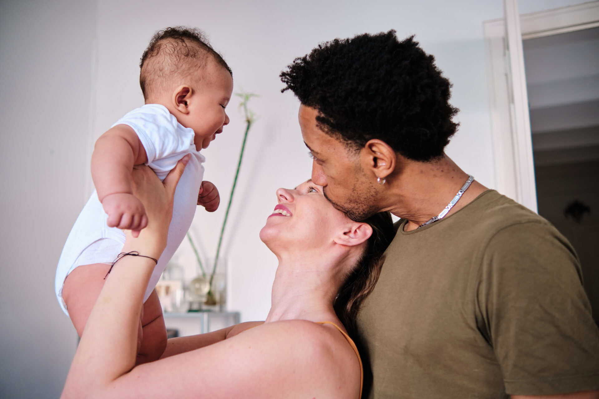 A couple joyfully holds up a laughing baby in a white onesie. The father tenderly kisses the mother's forehead, radiating warmth and family love.