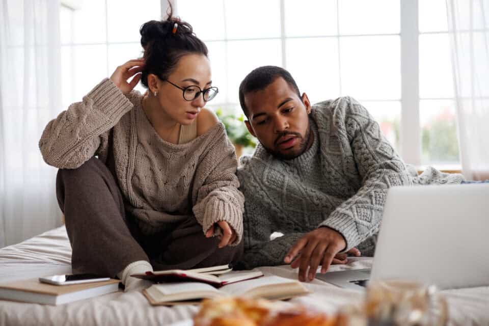 interracial couple in sweaters talking near books and laptop on bed at home