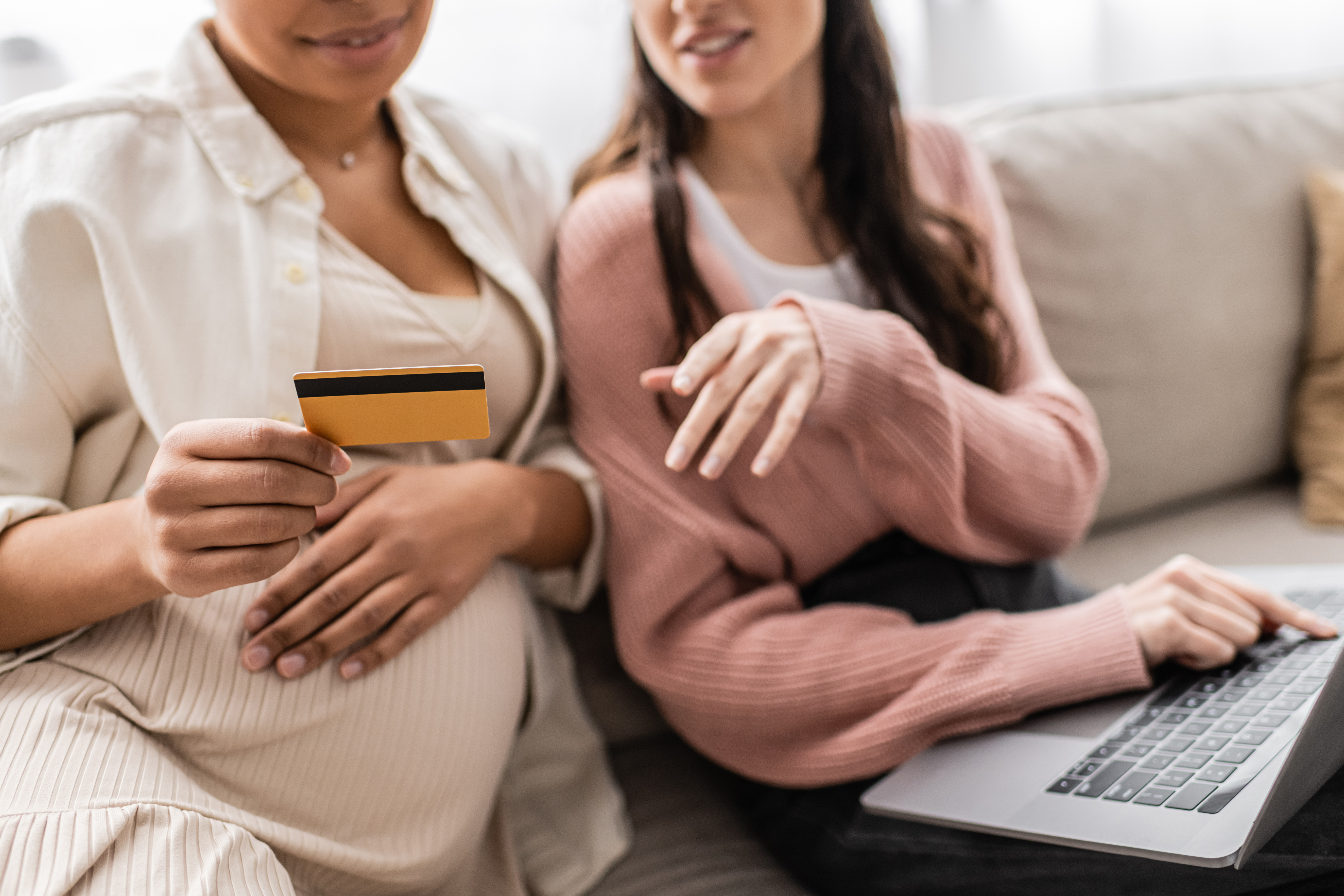 How Surrogate Pay Can Provide Meaningful Financial Support 1 Two women sitting on a couch, one holding a credit card, the other using a laptop. They appear focused, suggesting online shopping or payment.
