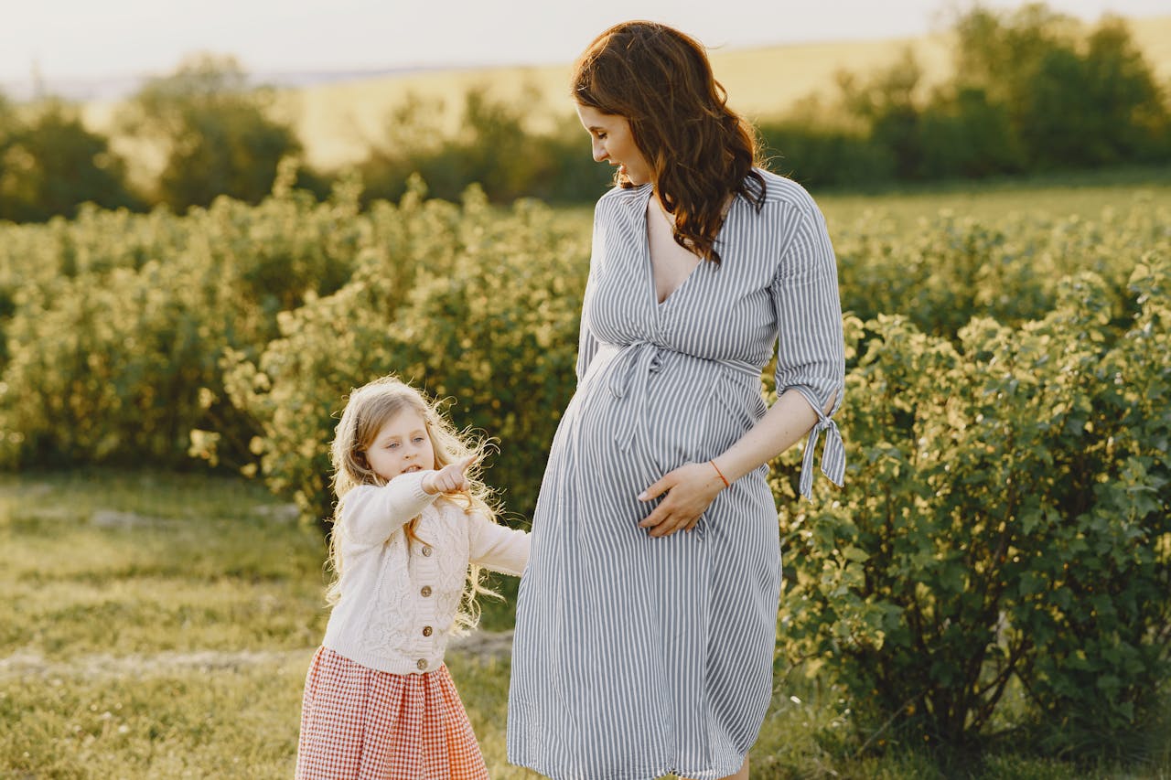 Pregnant woman in striped dress smiles at young girl in a field. The girl points ahead, both basking in warm sunlight, evoking a serene, joyful mood.